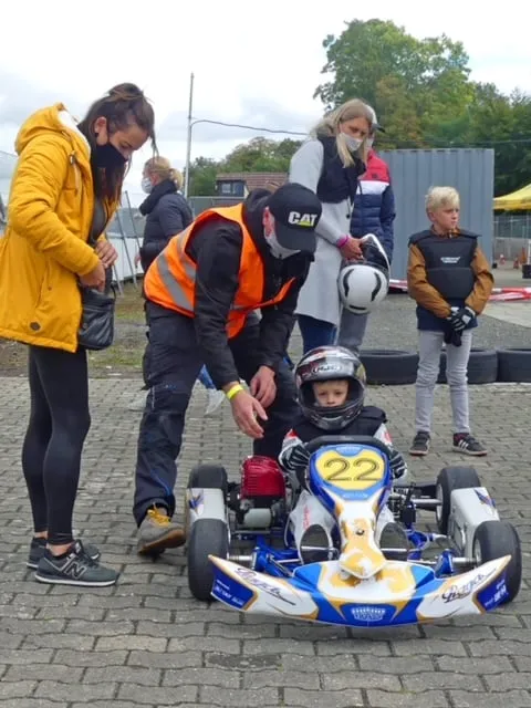 Ein Helfer in orangefarbener Warnweste unterstützt ein Kind im Kart im Fahrerlager.