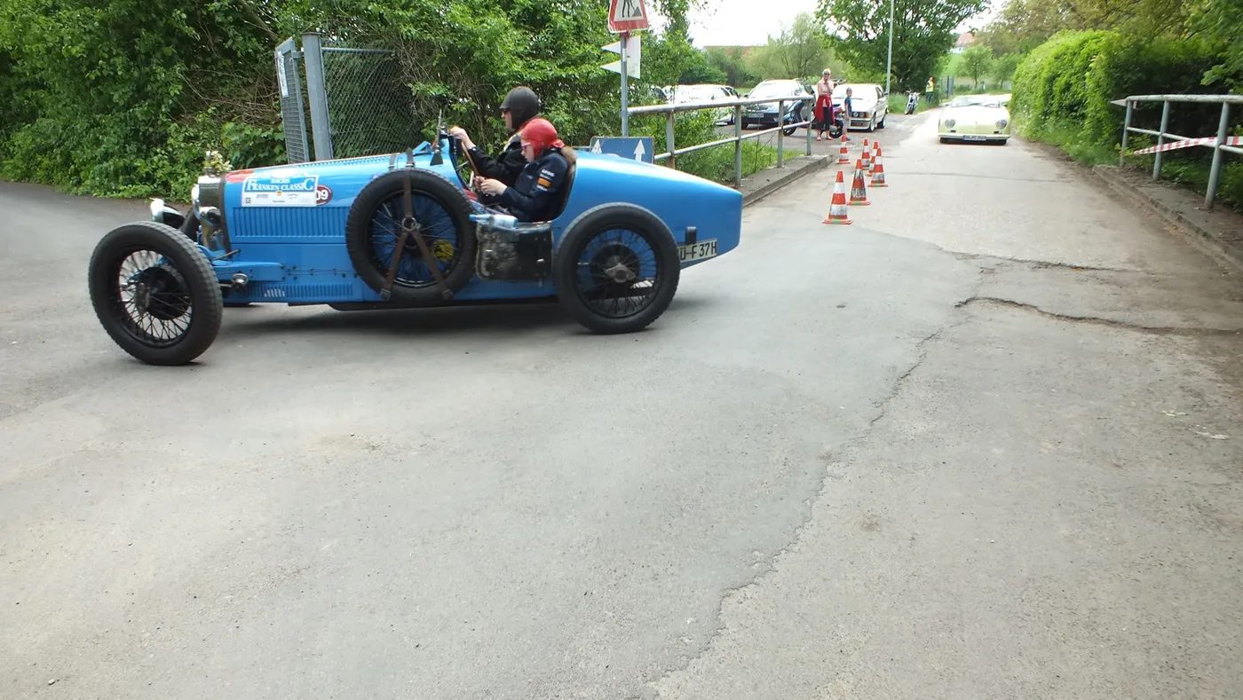 Blauer historischer Rennwagen mit Fahrer fährt durch eine Kurve, im Hintergrund Zuschauer.